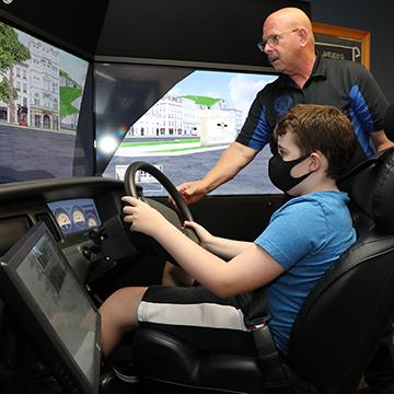 masked student sits in a driving simulator while an instructor stands behind him